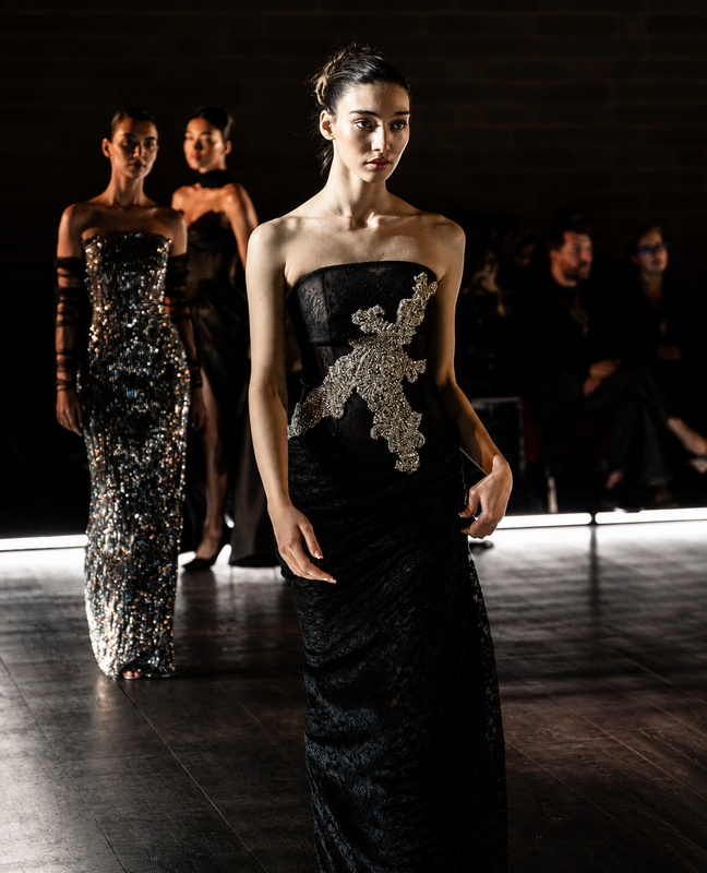 Black strapless evening gown with intricate silver embellishment on the bodice, presented on a fashion runway.